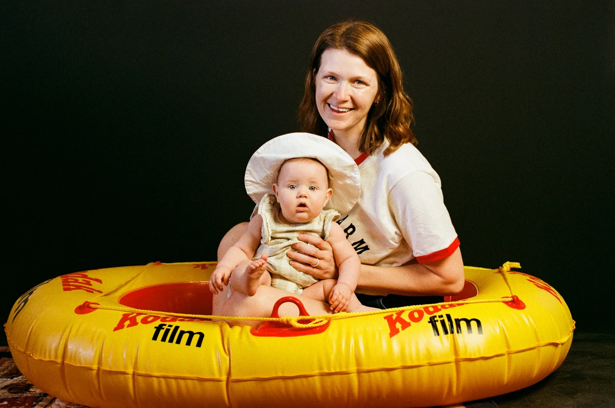 A smiling woman with brown hair holding a baby in a yellow KODAK film-themed inflatable boat against a dark background.