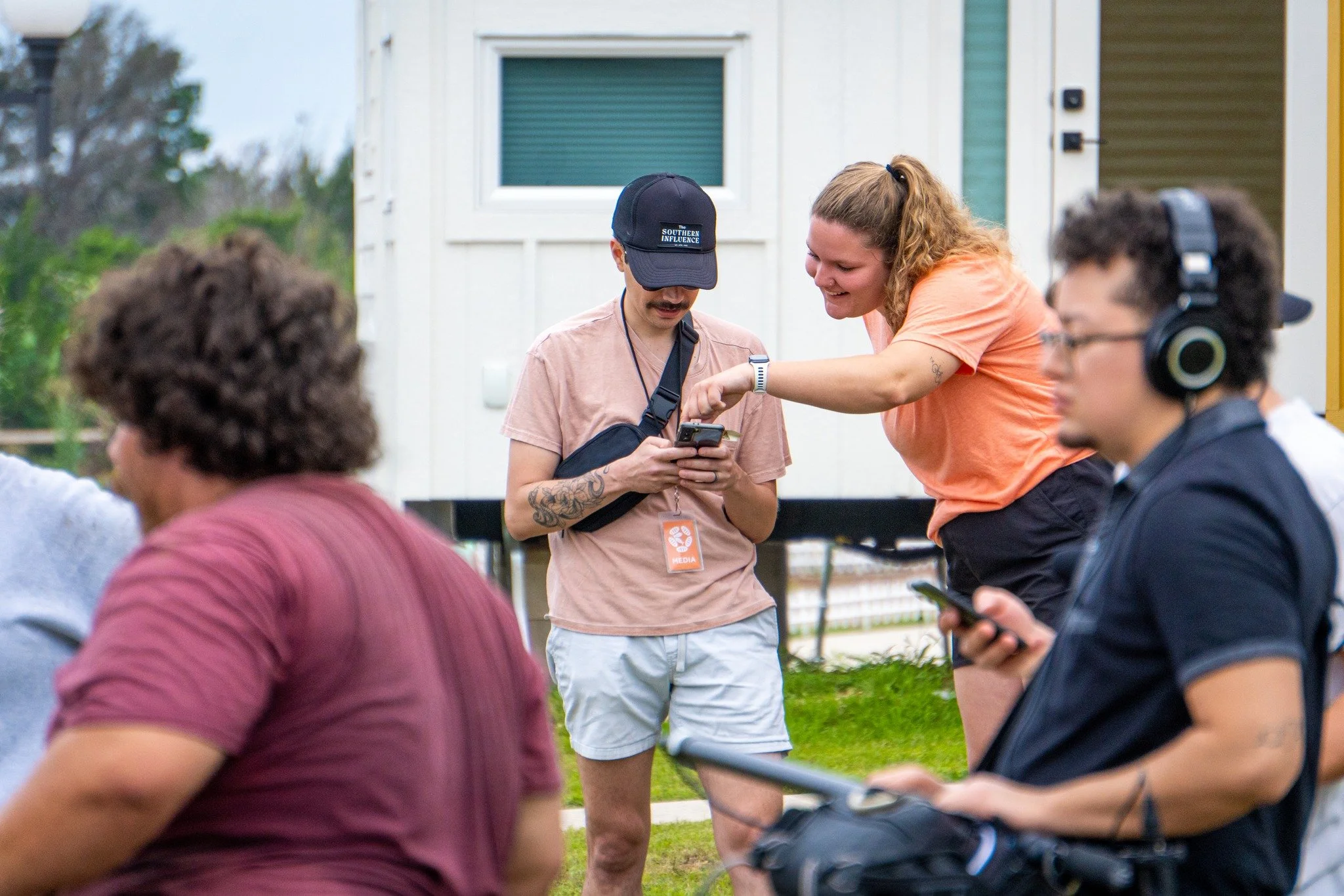 Group of people outdoors, one woman showing something on her phone to a man with a mustache and tattoos, others are using their phones and wearing headphones, with a house in the background.