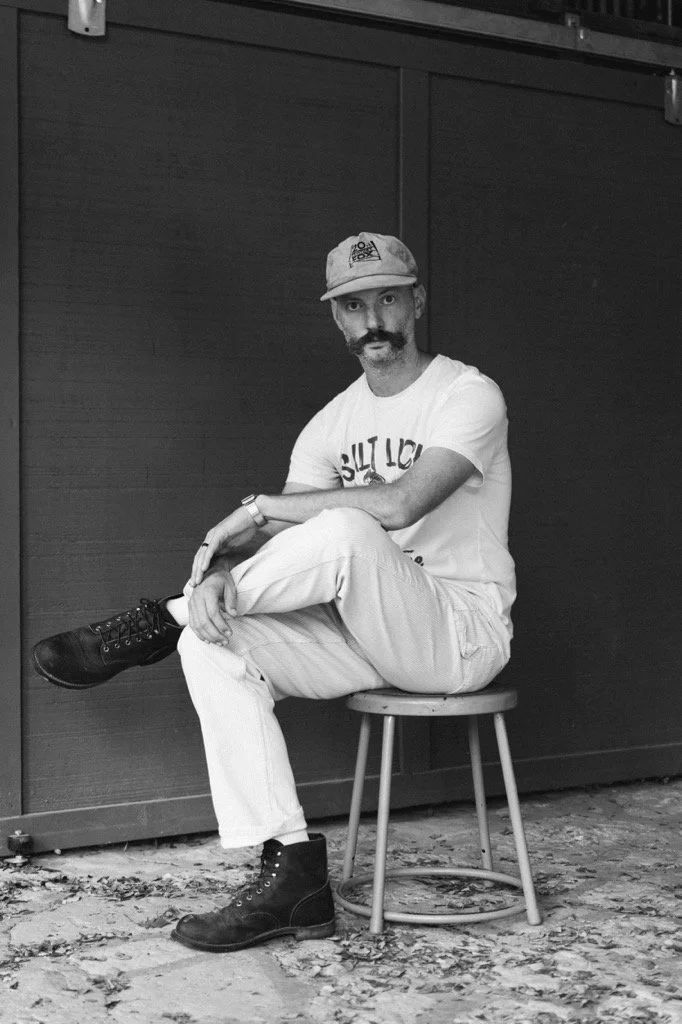 A man with a mustache and goatee, wearing a baseball cap, a white t-shirt, light-colored pants, and black boots, sitting on a stool against a dark wall, looking directly at the camera.