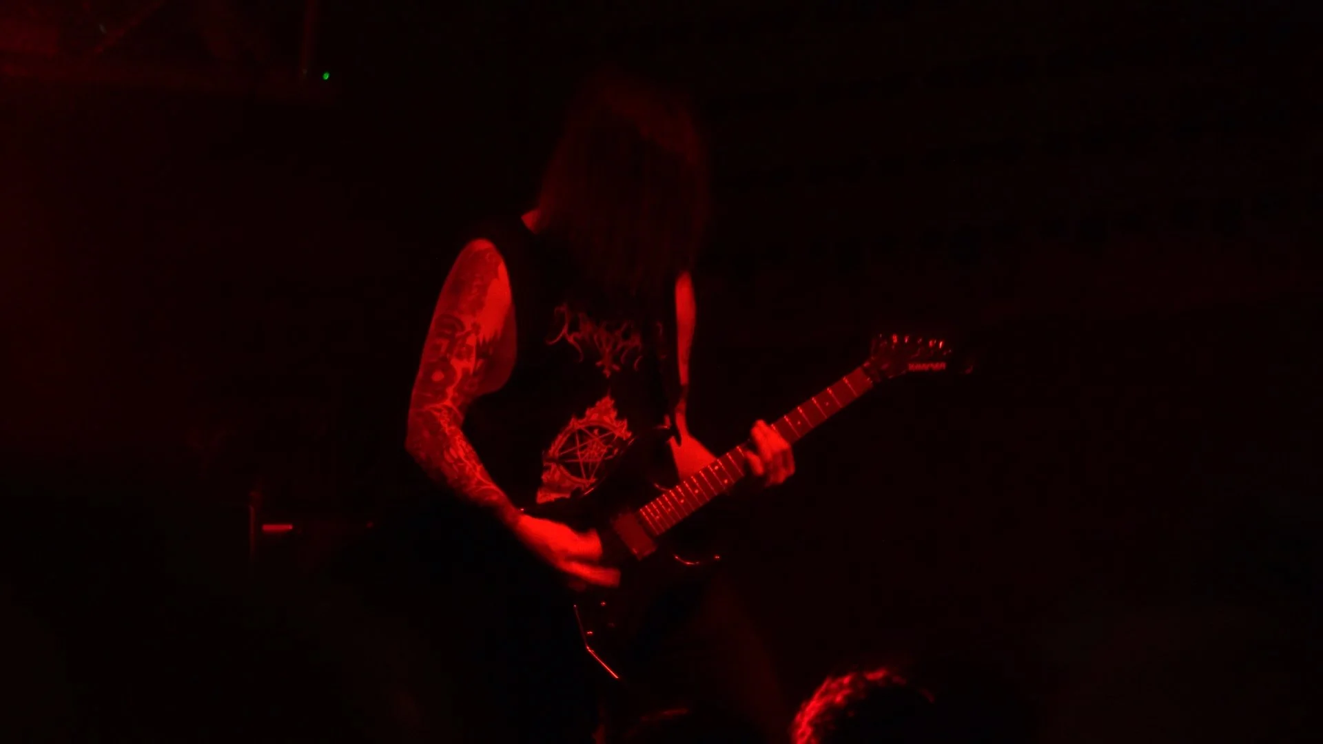 A person playing an electric guitar on stage, illuminated by red lighting, with long hair covering their face, wearing a sleeveless shirt with intricate designs and tattoos visible on their arm.