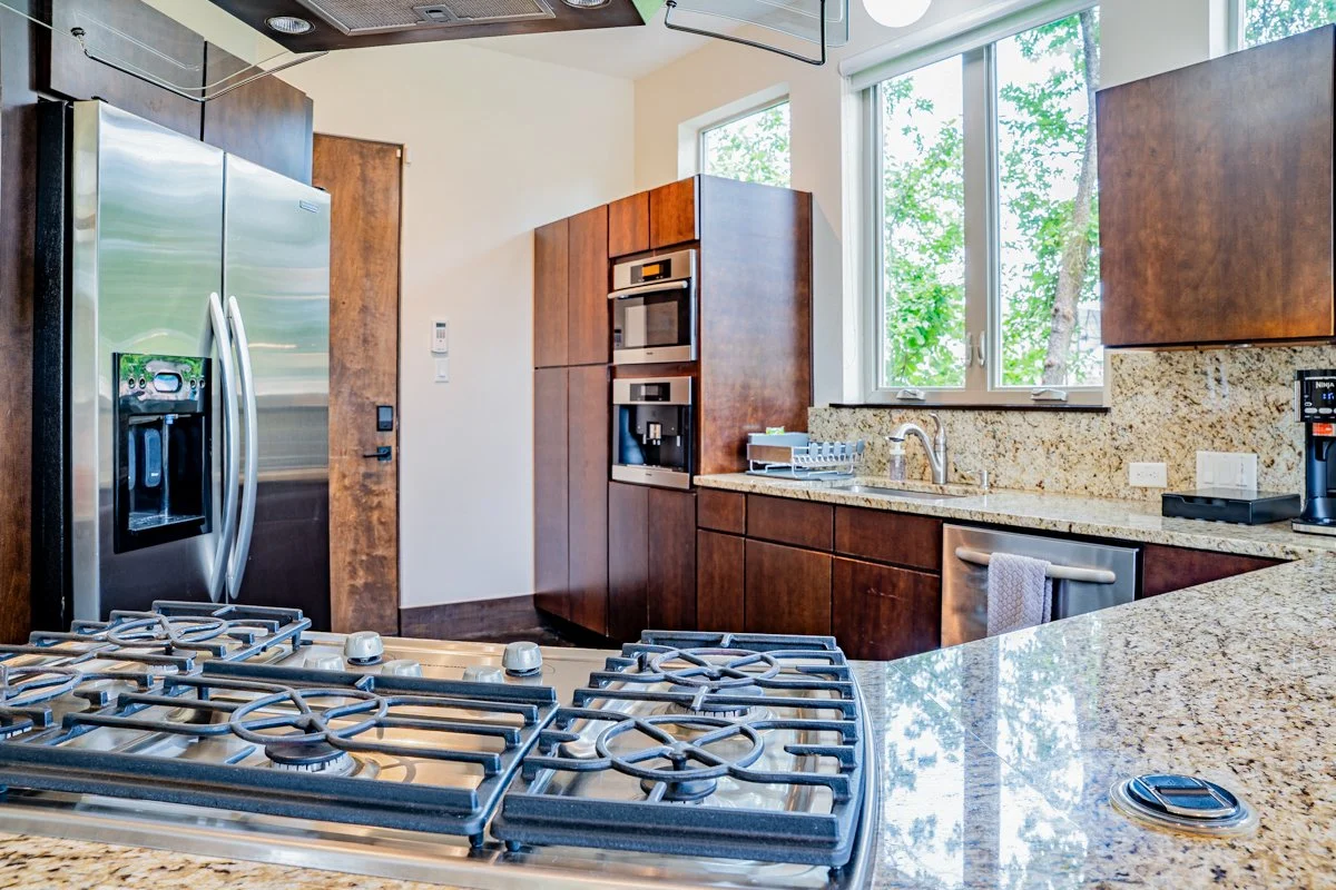 Modern kitchen with wooden cabinets, granite countertops, stainless steel refrigerator, gas stove, built-in microwave, coffee maker, and large window overlooking green trees.