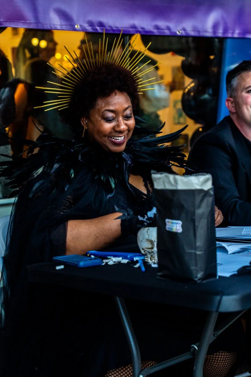 A woman wearing a black costume with feathered shoulders and a golden halo headpiece, smiling while sitting at a table.