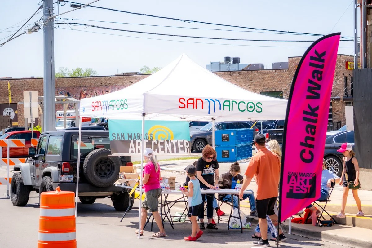 Children and adults gather around a table under a white canopy at the San Marcos Art Center during the San Marcos Art Fest, with a pink flag reading "Chalk Walk" nearby. A black Jeep with a spare tire on the back is parked behind the tent, and orange
