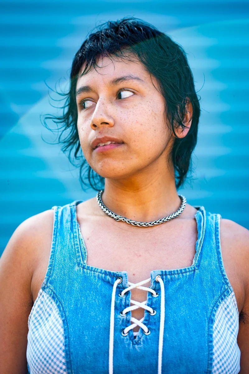 Young person with short dark hair and freckles wearing a denim sleeveless top with white laces and a silver chain necklace, standing in front of a water background.