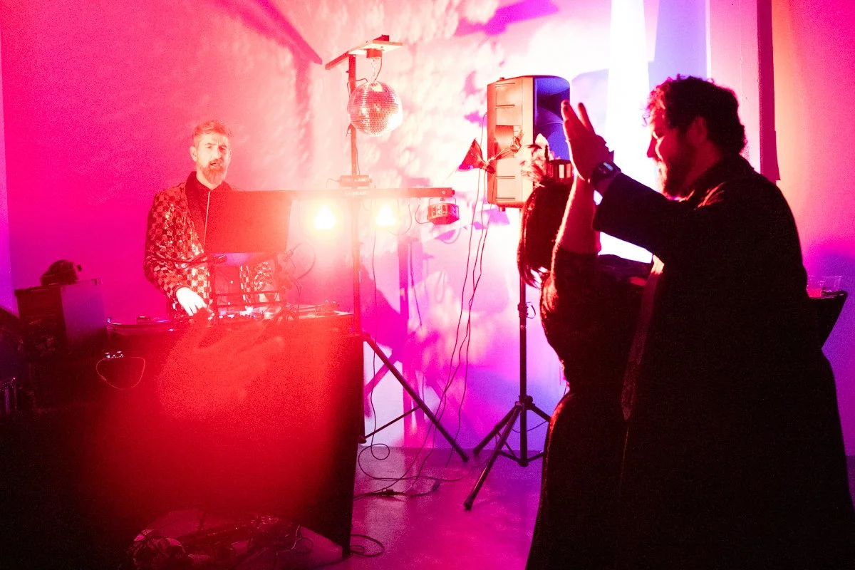 People dancing at a party with colorful pink and purple lighting, a DJ playing music, and a disco ball hanging from the ceiling.