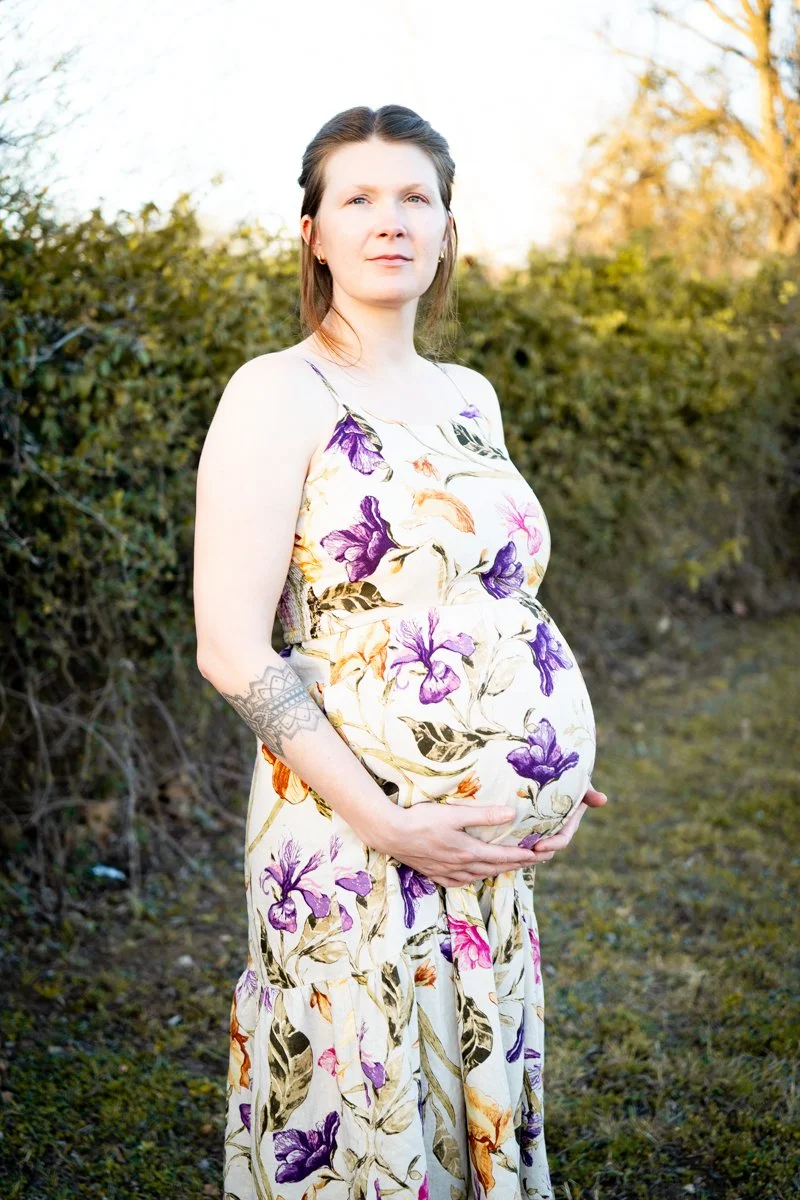 Pregnant woman standing outdoors in a floral dress, holding her belly with both hands, with bushes and trees in the background.