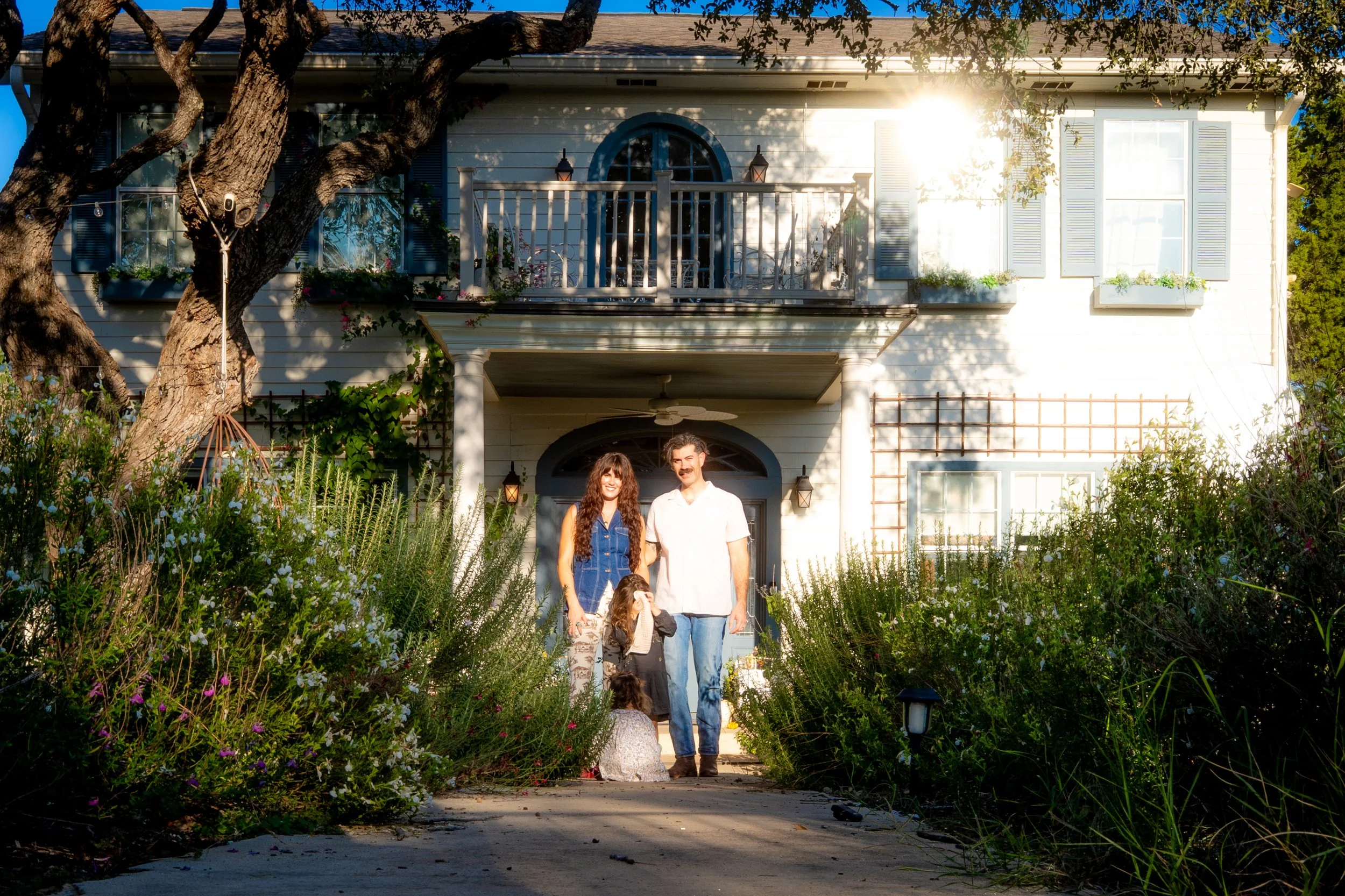 A family of four standing in front of a white house with a porch, surrounded by lush garden plants, with sunlight shining from the right side.