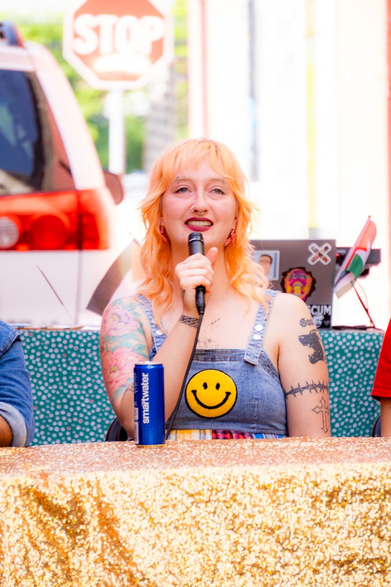 A woman with orange hair and tattoos, sitting at a table with a golden glitter tablecloth, speaking into a microphone, with a STOP sign and colorful background behind her.