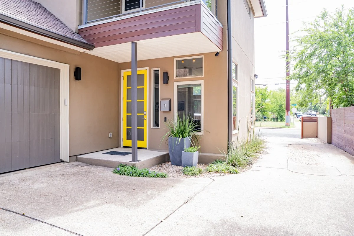 Exterior of a modern apartment building with a yellow front door, potted plants, a mailbox, and large windows, with a parking and street visible in the background.