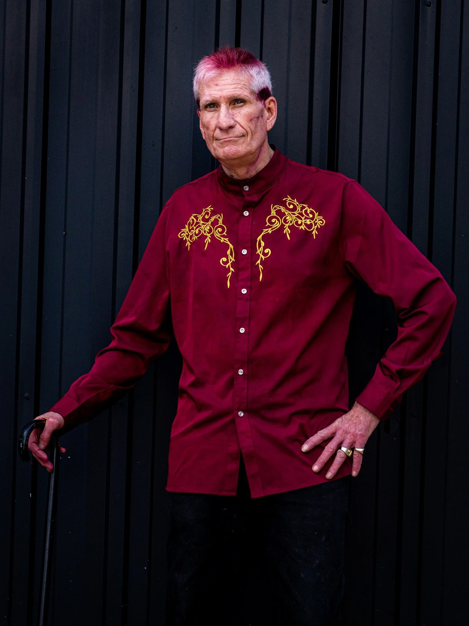 An older man with dyed pink and white hair, wearing a maroon shirt with yellow embroidery, standing against a black paneled wall, holding a cane with his left hand, and has his right hand on his hip.