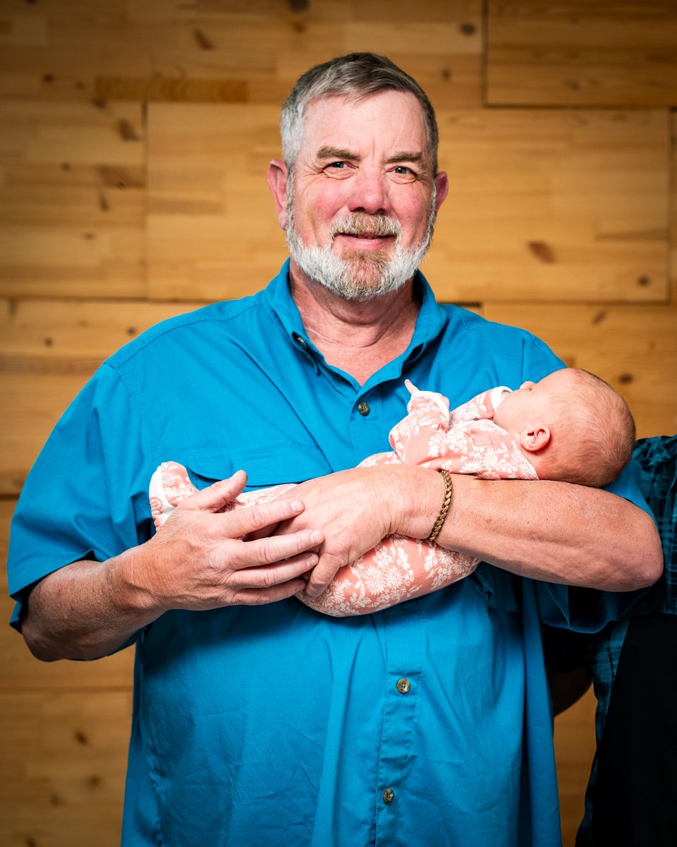 An elderly man with gray hair and a beard, wearing a blue button-up shirt, holding a newborn baby wrapped in a pink and white blanket, standing in front of a wooden paneled wall.