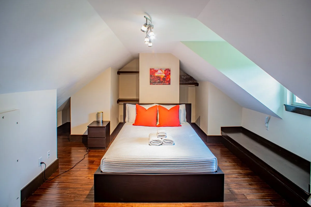 A cozy bedroom with a sloped ceiling, a queen-sized bed with white linens and two orange pillows, a small bedside table, and a window with natural light.
