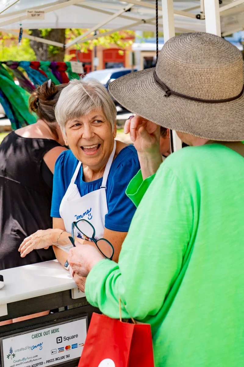A smiling older woman with short gray hair, wearing a blue shirt and apron, talking to a person in a green shirt and large sun hat at an outdoor market stall. The stall has colorful textiles hanging in the background.