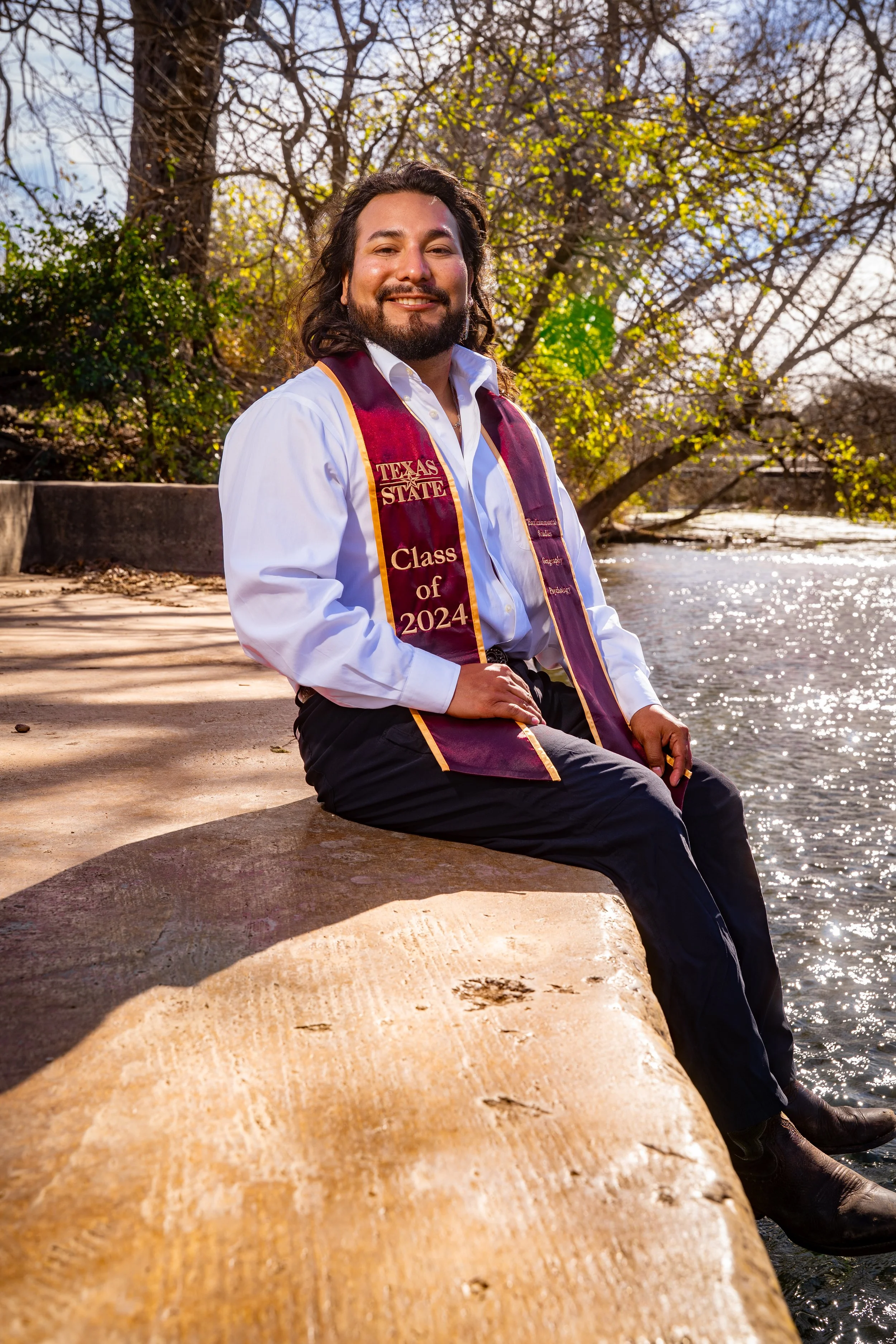A young man in white dress shirt and dark pants sitting on a stone ledge by a river, wearing a maroon graduation stole that reads "Texas State, Class of 2024." He is outdoors with trees and sunlight in the background.