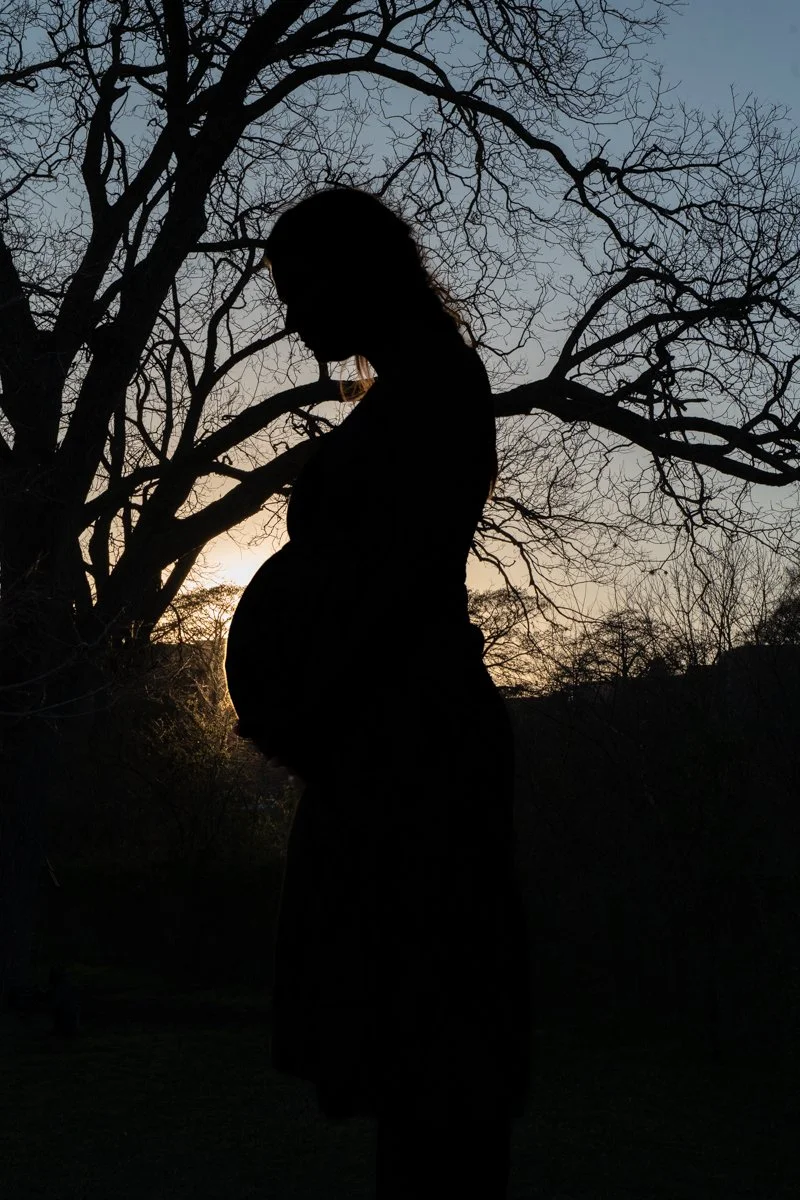 Silhouette of a pregnant woman standing outdoors at sunset or sunrise, with leafless trees in the background.