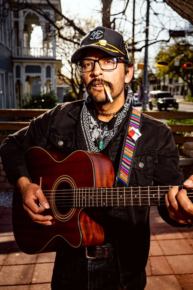 A man with glasses, a cigarette in his mouth, and a black cap playing an acoustic guitar outdoors in a neighborhood with trees and houses in the background.