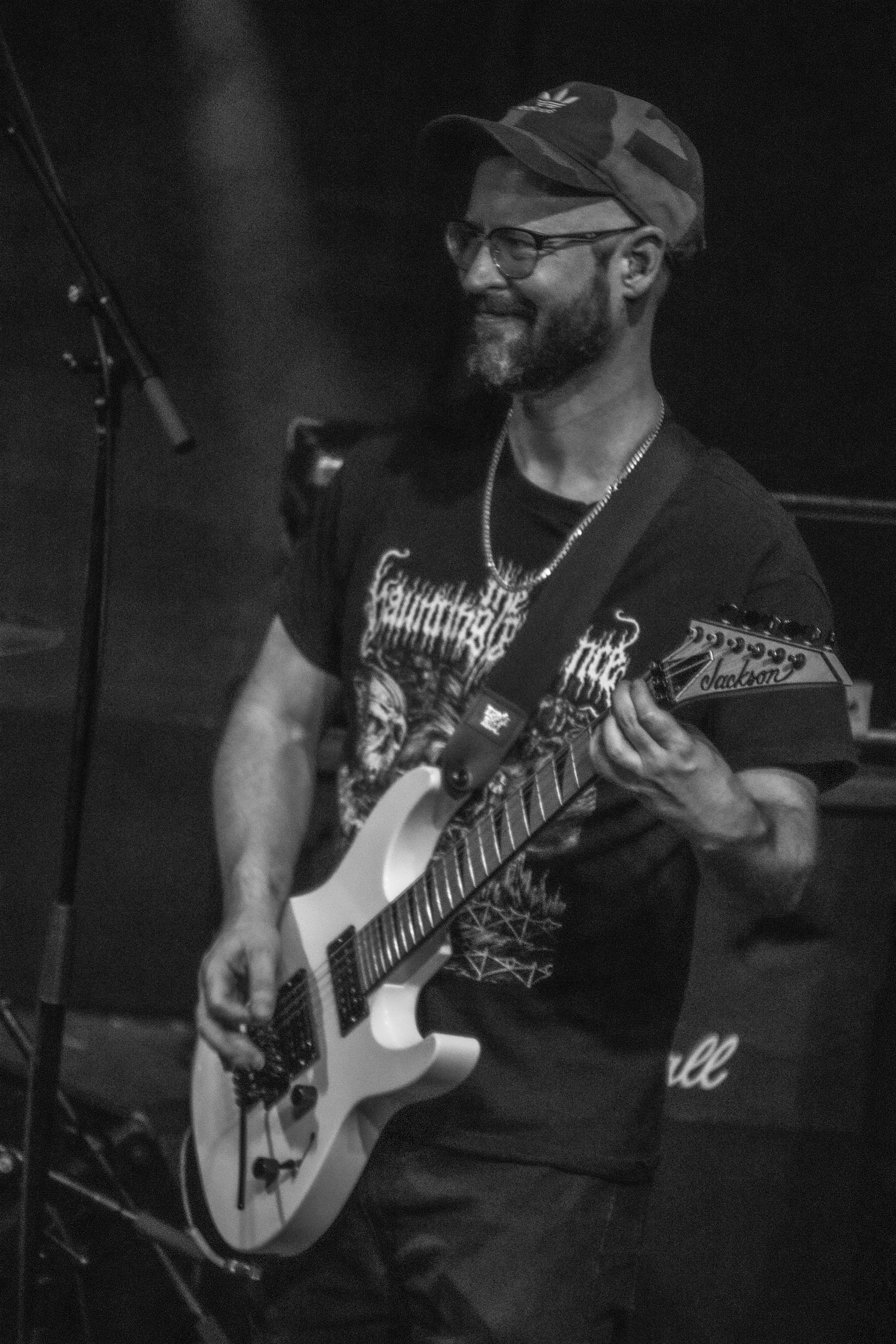 A man with glasses, a beard, and a camouflage cap playing an electric guitar on stage, smiling, with a microphone stand nearby.