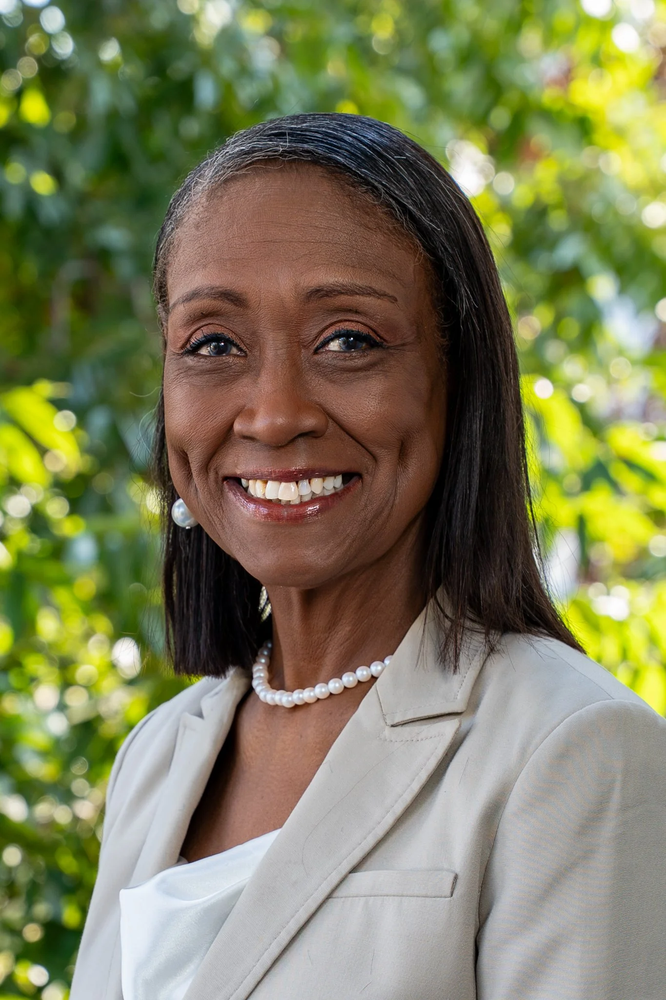 A smiling African American woman with dark hair, wearing pearl earrings, a pearl necklace, and a beige blazer, outdoors with green foliage in the background.