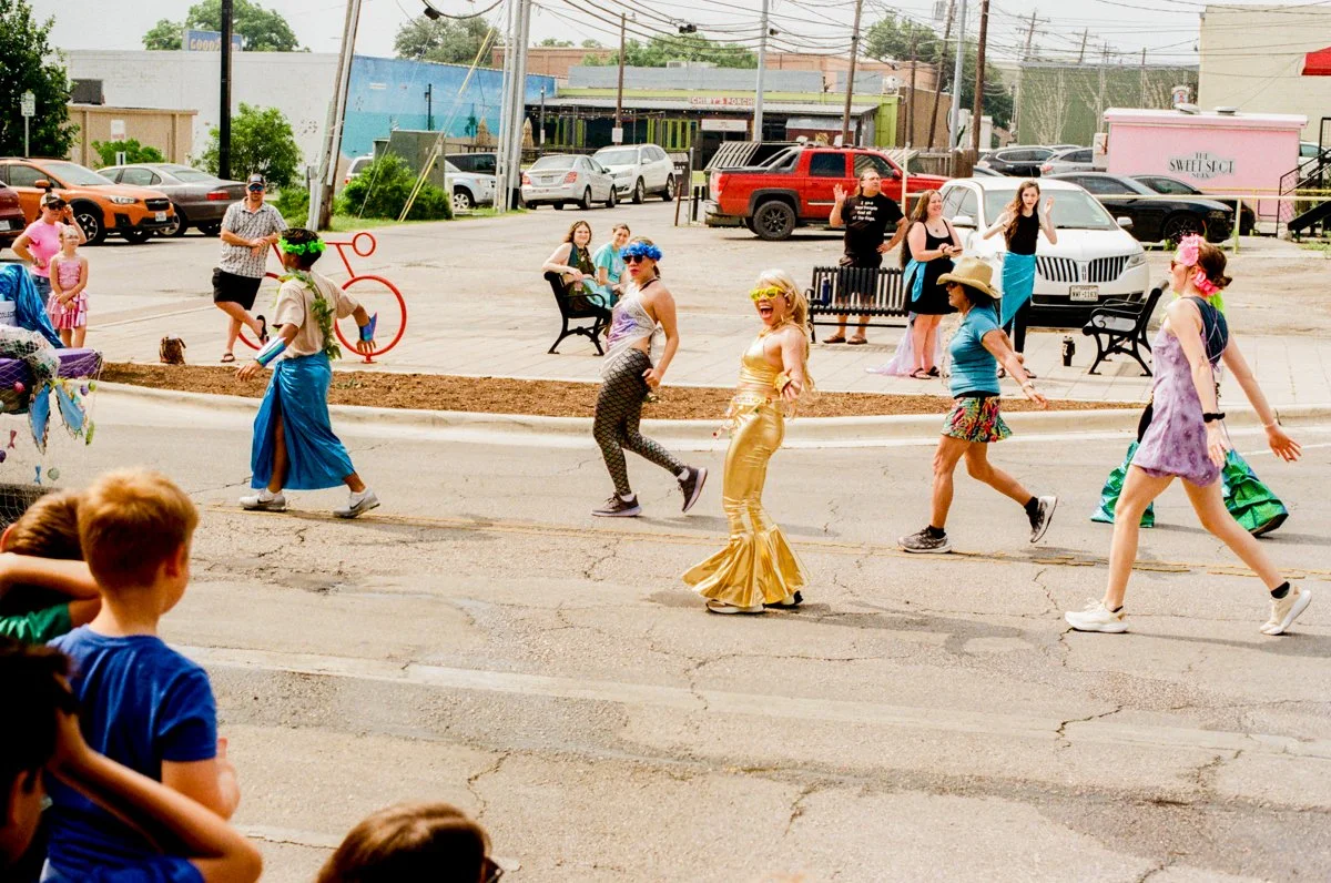 People dressed in colorful costumes walking and dancing across a street during a parade or festival, with spectators watching from the sidewalk.