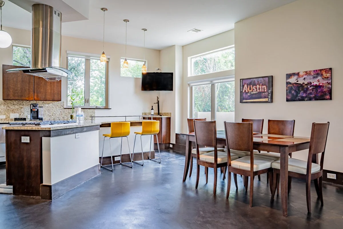 Open-concept kitchen and dining area with large windows, a stainless steel range hood, granite countertops, a small breakfast bar with two yellow chairs, a wall-mounted TV, and wood dining table with six chairs. Wall art includes two colorful framed 