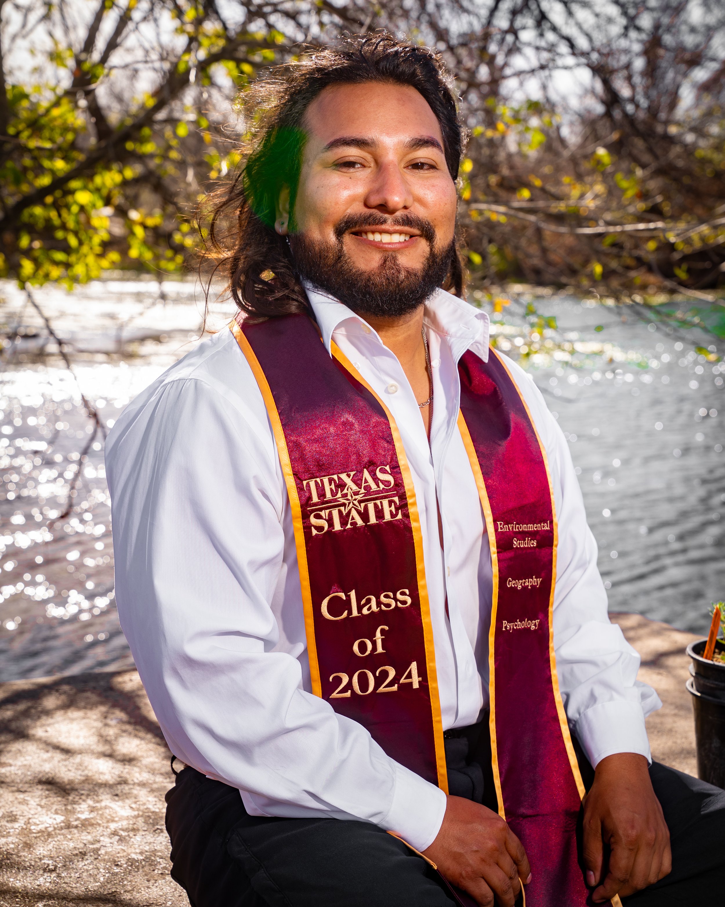 A man with long dark hair and a beard, smiling, wearing a white shirt and a maroon graduation stole with gold edges that reads 'Texas State Class of 2024' and lists environmental studies, geography, and psychology, sitting outdoors near a river with 