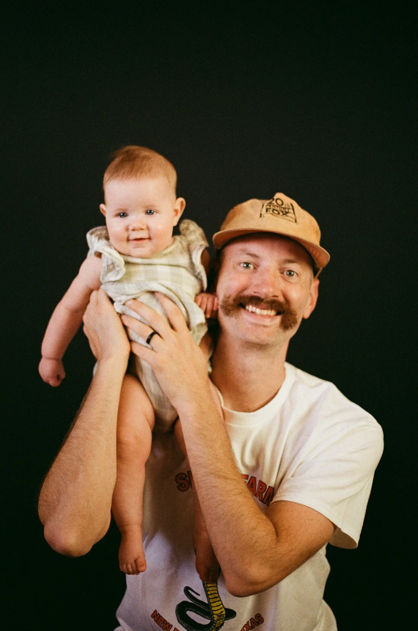 A man with a mustache, smiling, holding a young child on his shoulder. The man is wearing a tan cap and a white t-shirt, and the child has short hair and is wearing a light-colored sleeveless outfit. Both are against a dark background.
