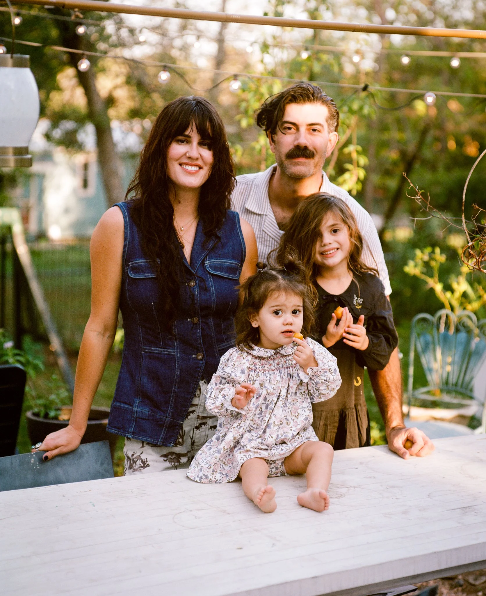 A family of four enjoying an outdoor gathering in a backyard during the evening. The mother stands to the left, wearing a sleeveless denim vest, and the father is to the right with a mustache and a short-sleeved shirt. Two young girls are in front of