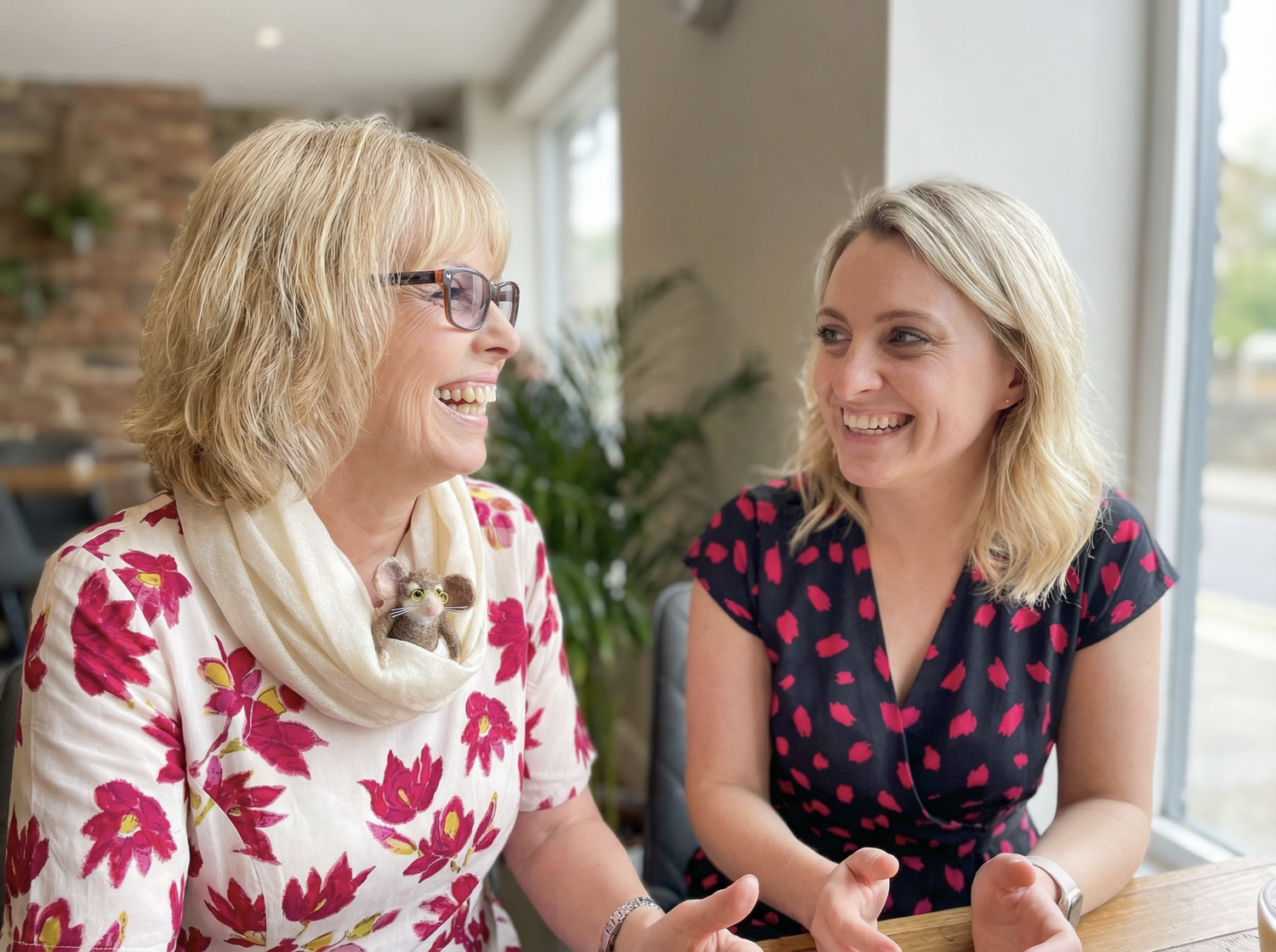 Two women laughing and talking at a table in a bright, cozy cafe. One woman is wearing glasses and a floral top with a felted mouse in her scarf, and the other woman is wearing a black dress with pink heart patterns.