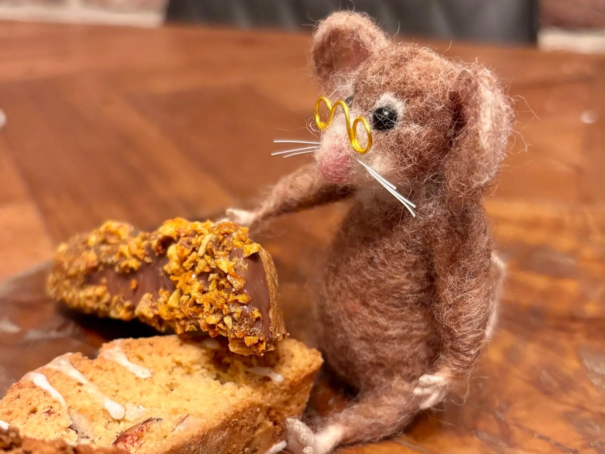 A small felted mouse figurine with glasses and white whiskers sitting on a wooden table, reaching over to a plate of biscotti.