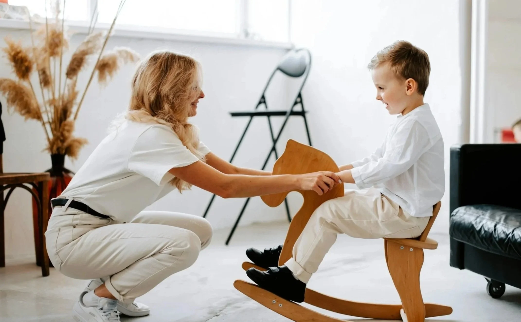 A woman playing with a young boy on a wooden rocking horse in a bright room, both smiling and holding hands.