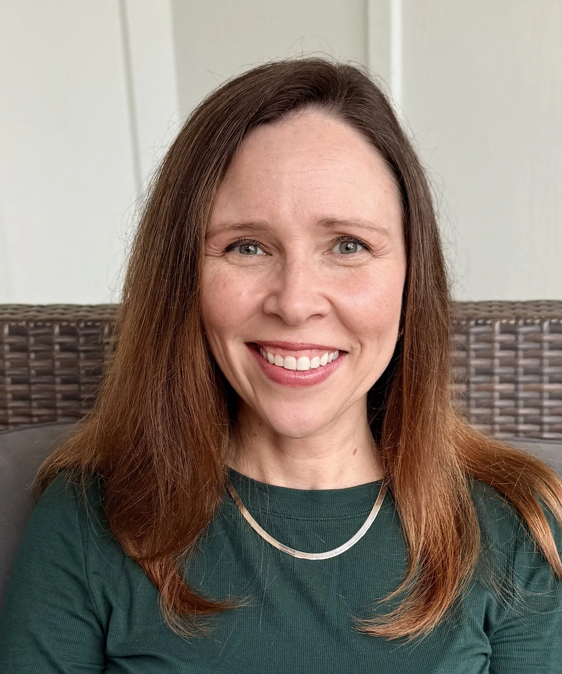 A woman with long reddish-brown hair, smiling, wearing a dark green top and a gold necklace, sitting on a wicker chair in a neutral space.