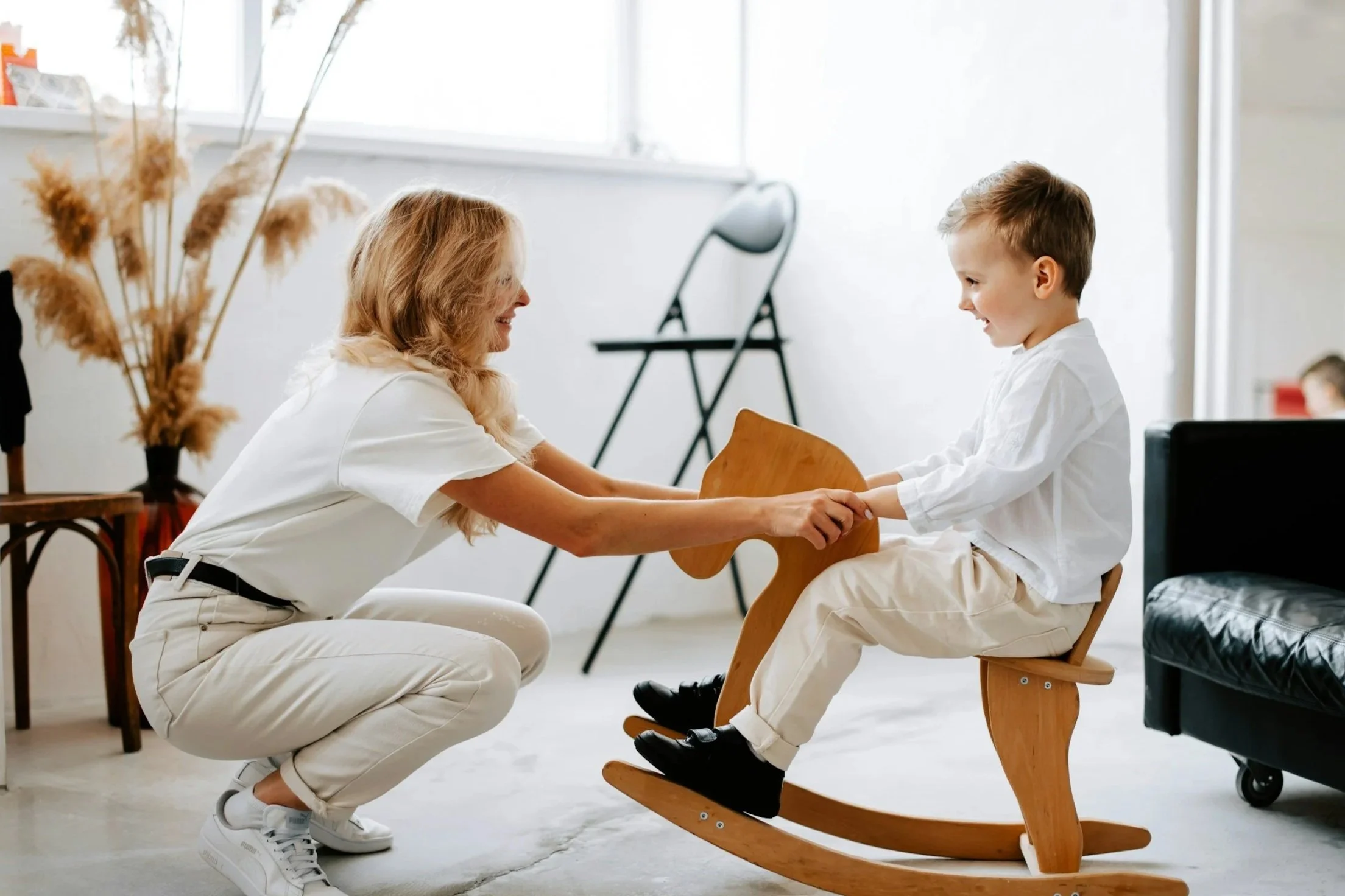 A woman kneeling on the floor reaching out to a young boy sitting in a wooden rocking chair, holding his hands, in a bright room with minimal decor