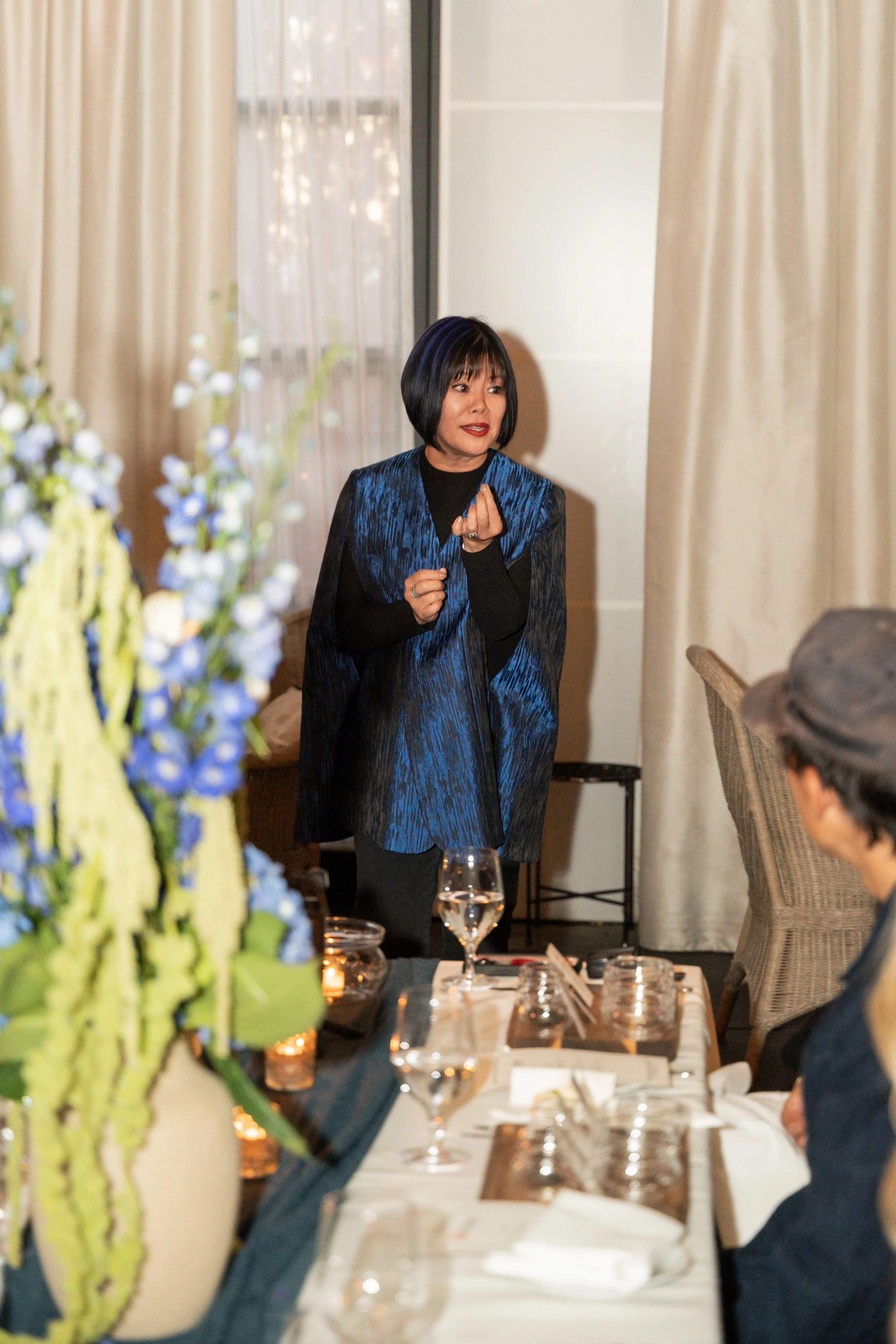 Woman standing and speaking at a formal event, with floral arrangements and glasses of wine on the table in front of her.