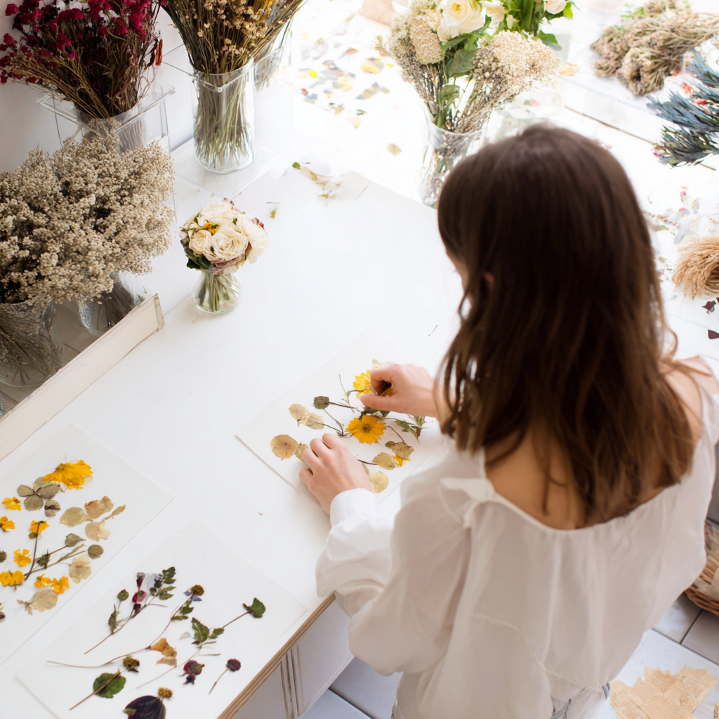 A woman arranging preserved flowers on a white table, surrounded by various vases of dried and fresh flowers, with floral pressed flowers on the table.