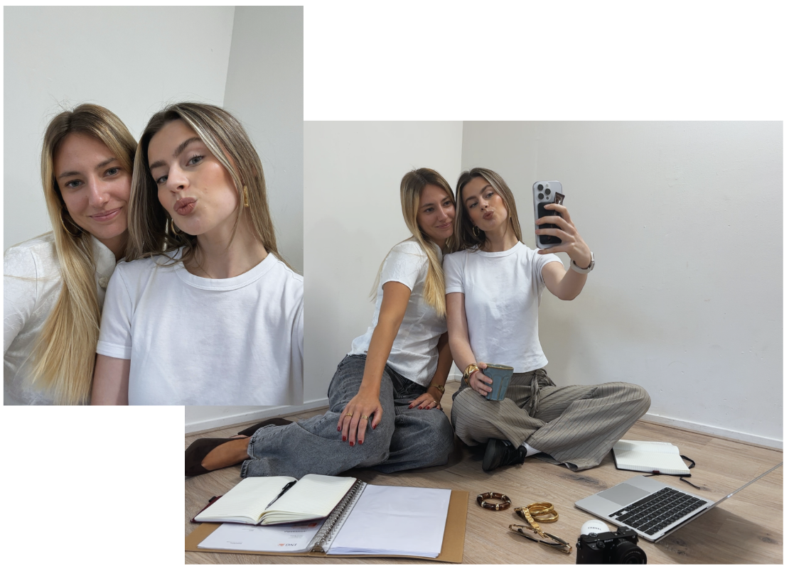 Two young women taking a mirror selfie while sitting on the floor. There is an open notebook, some folders, jewelry, a camera, and a laptop on the floor.