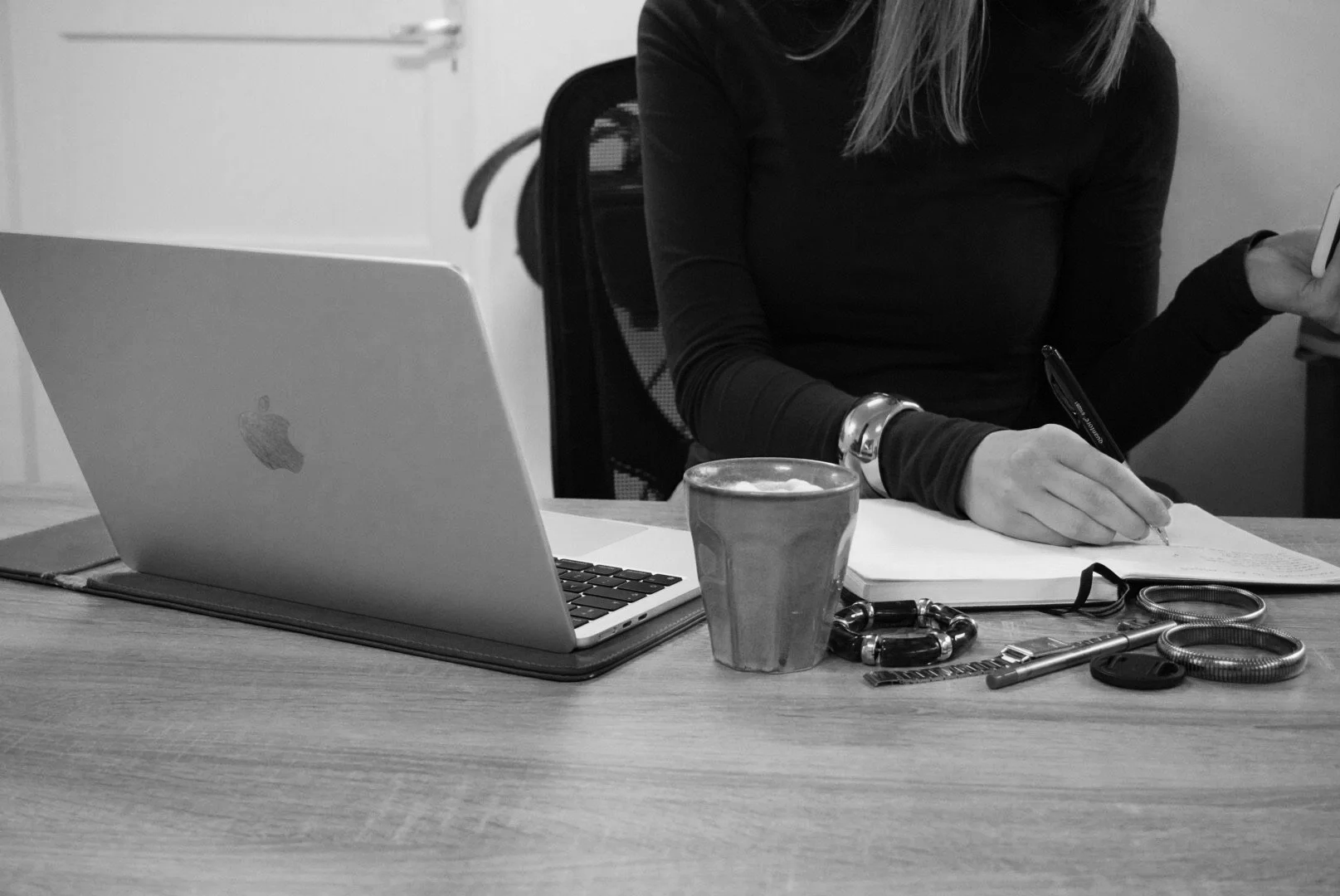 Woman sitting at a desk writing in a notebook with a laptop, cup, and various jewelry items on the desk in black and white.