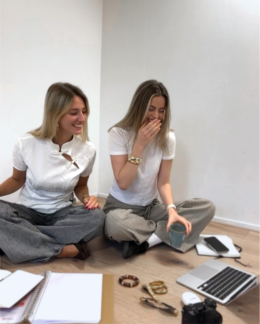 Two young women sitting on a floor with laptops, notebooks, and accessories, laughing and chatting, in a casual office setting.