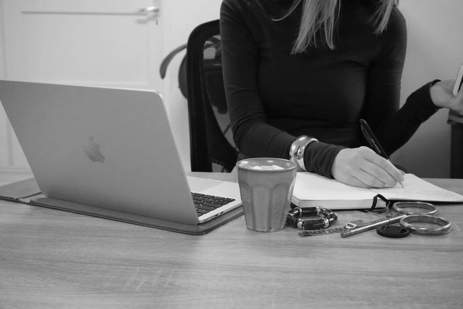 Person sitting at a desk with a laptop, taking notes with a pen, and a cup of coffee, with various personal belongings on the table.