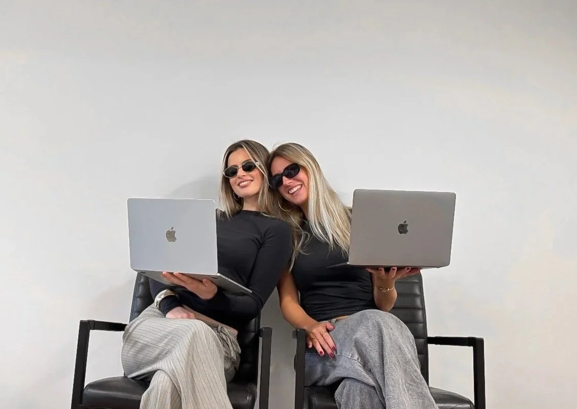 Two women sitting on black chairs, holding laptops, wearing black tops and sunglasses, smiling at the camera.