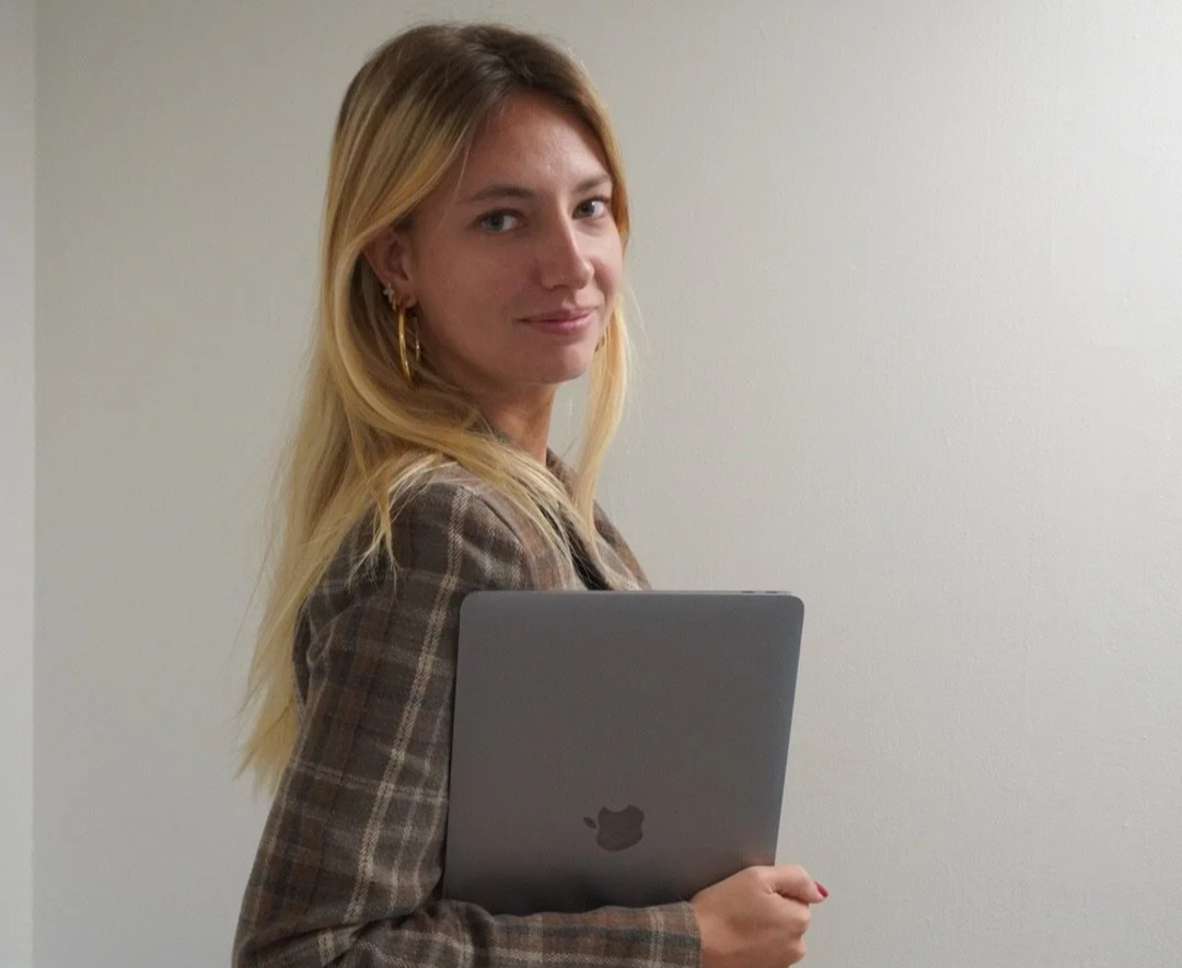 A woman with long blonde hair holding a silver laptop and looking at the camera.