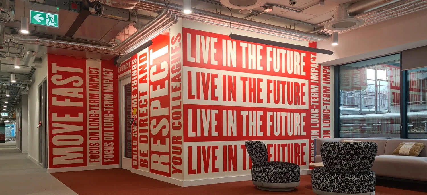 Interior office lounge area with red and white wall murals displaying motivational and inspirational messages, including 'MOVE FAST', 'BE RESPECTFUL', and 'LIVE IN THE FUTURE'. There are two patterned chairs, a beige sofa, and large window with a vie