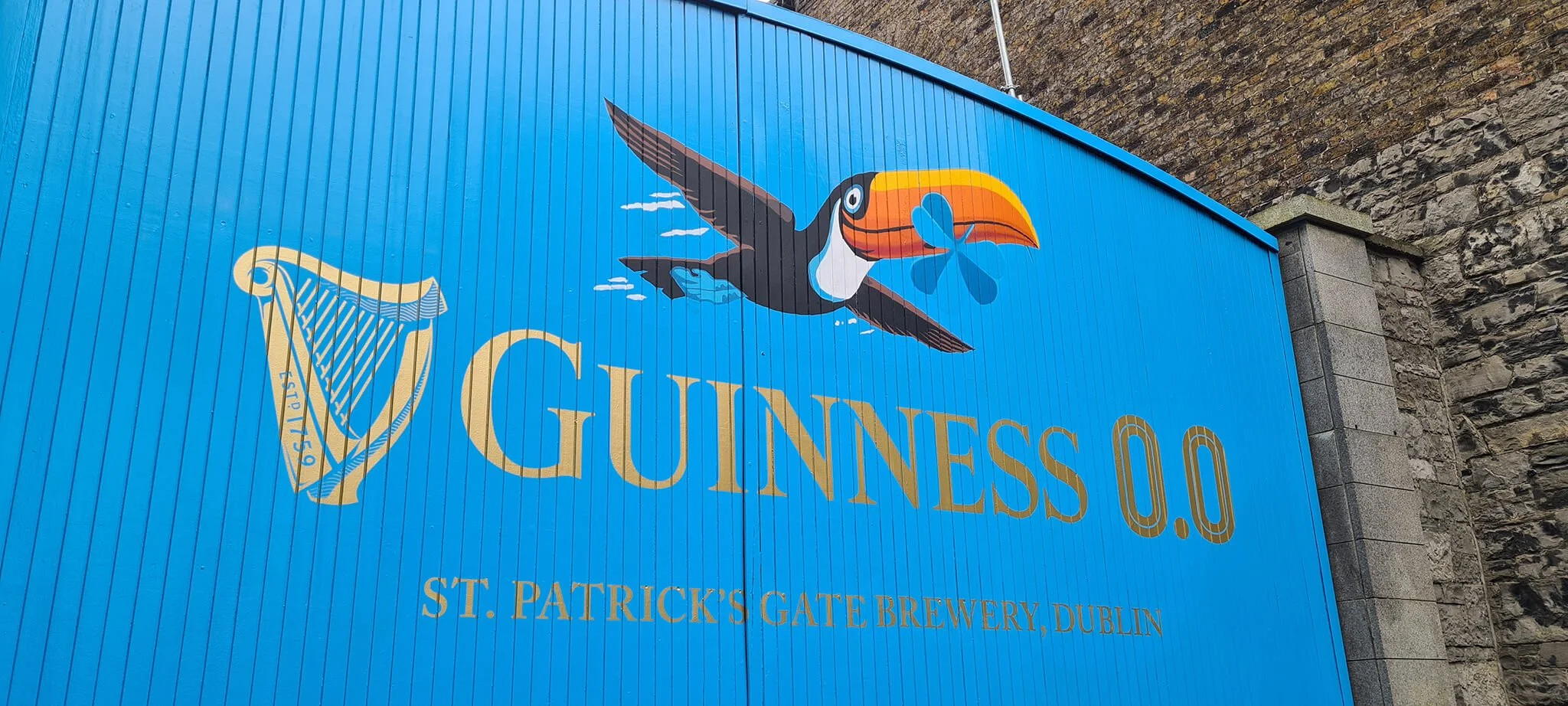 Blue sign with the Guinness logo, featuring a colorful toucan holding a blue shamrock in its beak, and the text "Guinness 0.0" and "St. Patrick's Gate Brewery, Dublin".