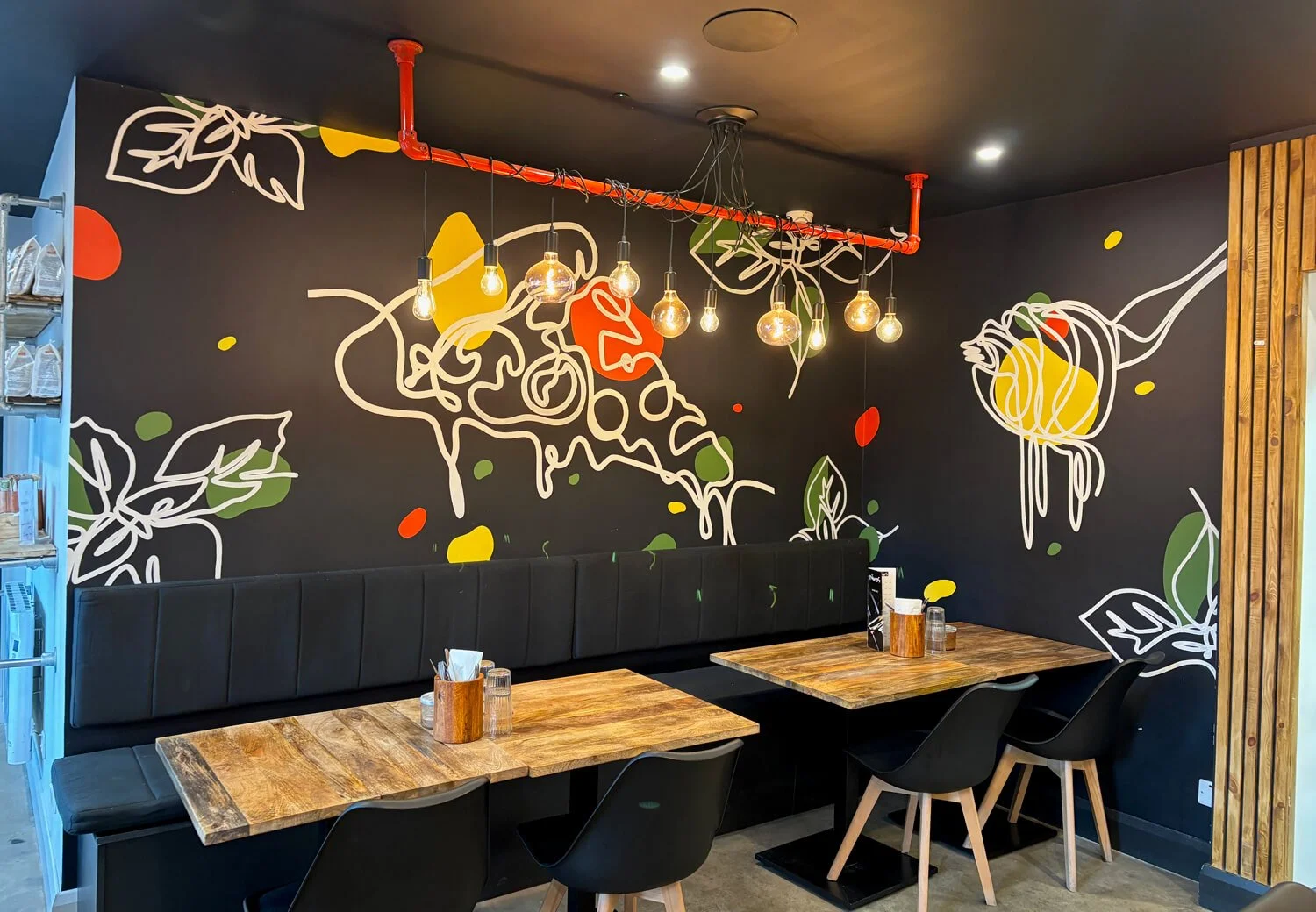 Interior mural in an office building featuring a black wall with a colourful floral mural, two wooden tables with black chairs, a long black padded bench, and hanging pendant lights with exposed bulbs.