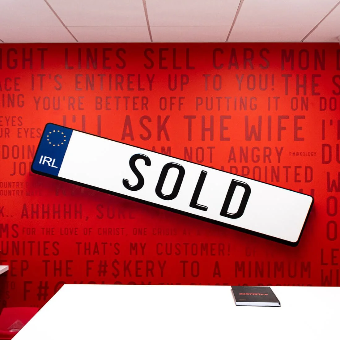 A European license plate with the word 'SOLD' on a white background and a blue section with yellow stars, positioned diagonally in front of a red wall with various phrases and text in black and red.