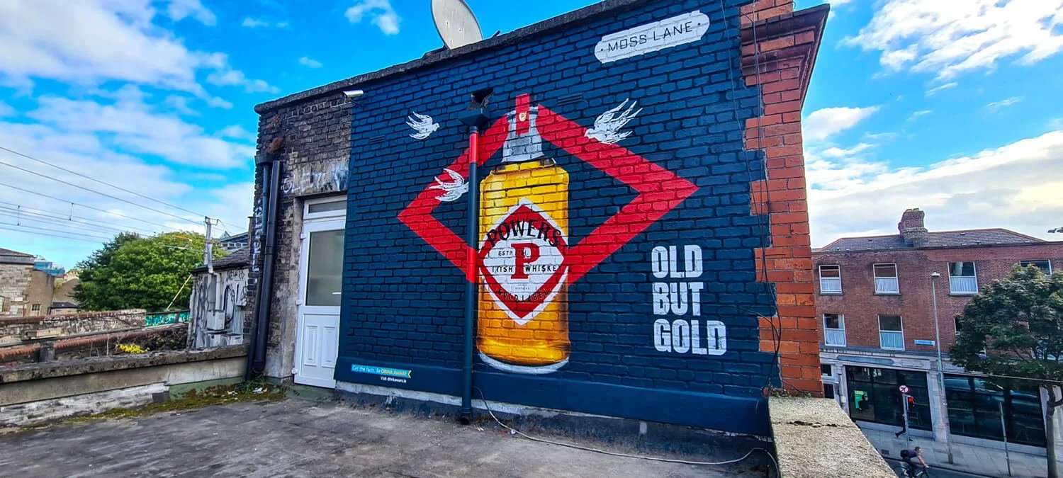 A colourful mural painted on the side of a brick building, featuring a large bottle of Powers whiskey with the slogan 'Old But Gold' next to it. 