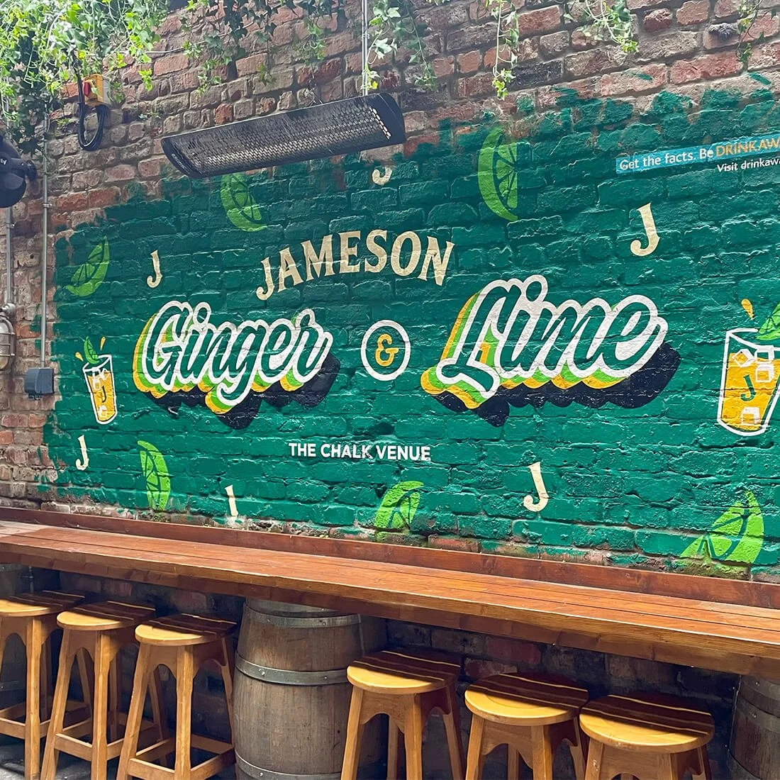 Decorative brick wall with a mural advertising Jameson Ginger & Lime cocktails at The Chalk Venue. The mural features illustrations of lime wedges and drink glasses with straws, along with the text 'Jameson Ginger & Lime' and some letters 'J' and 'J' scattered around. There's a long wooden bar counter with stools below the mural.