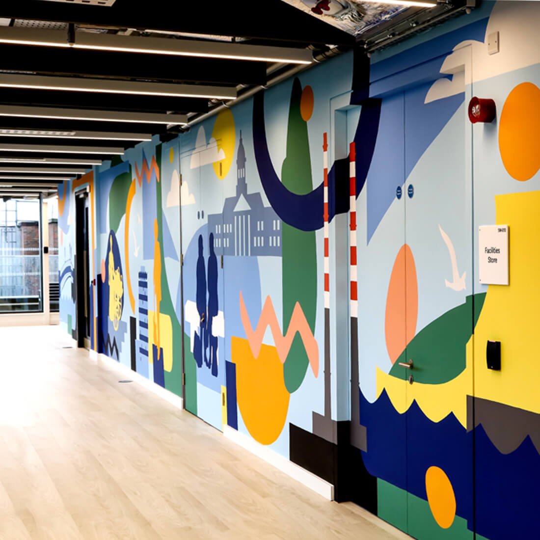 Colorful mural of abstract shapes, silhouettes, and cityscape scenes on a hallway wall. The mural features bright colors including blue, yellow, orange, green, and black, with a sign indicating facilities store.