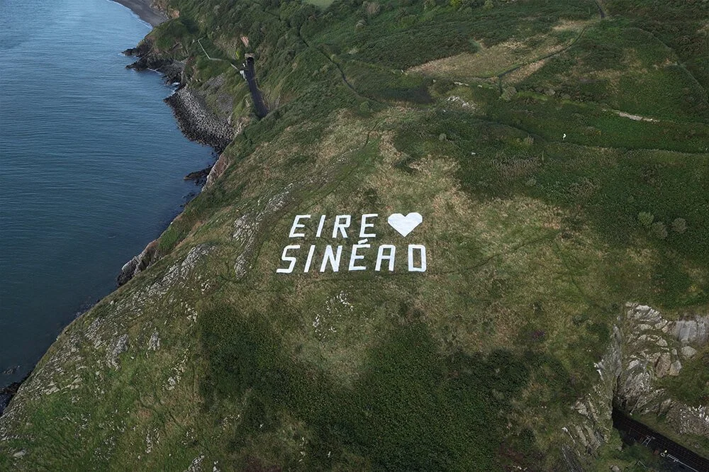 A hillside with large white letters spelling 'EIRE' and 'SINEAD' separated by a white heart symbol, overlooking a body of water with a coastline and green forested landscape.