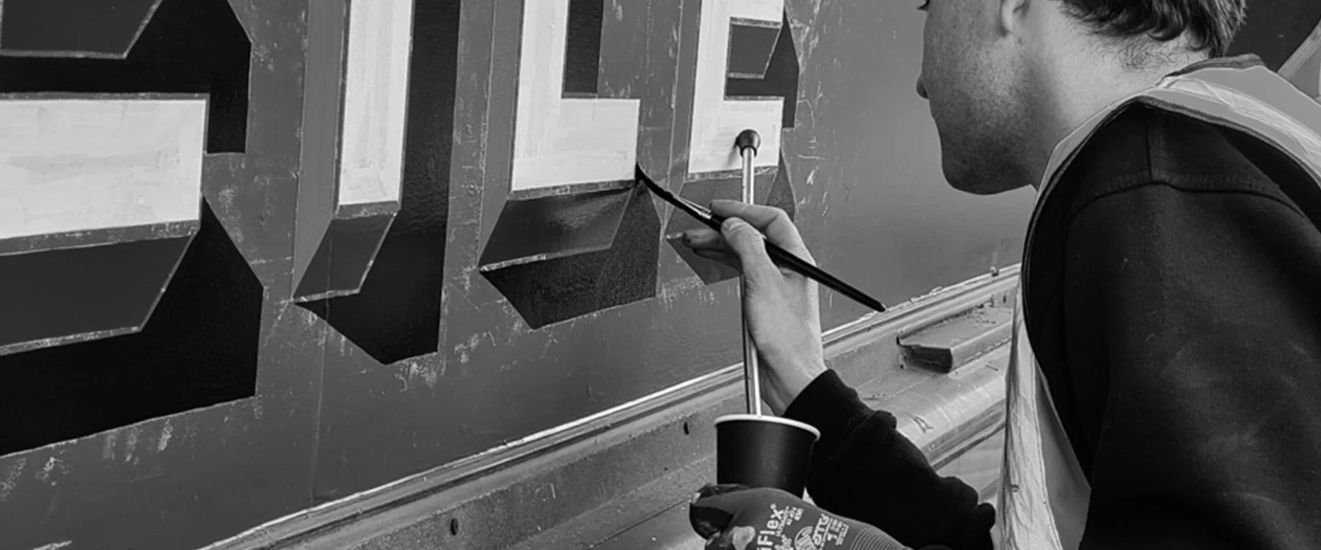 A person painting 3D lettering on a wall with a paintbrush, using a cup of paint and wearing gloves.