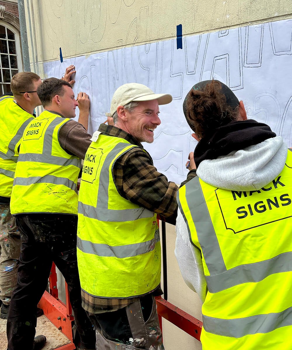 Group of people wearing high-visibility vests labeled 'Mack Signs' creating a large outdoor sign or mural on a wall.